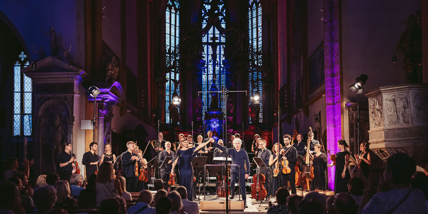 Konzertszene in einer prunkvollen Kirche mit hochaufragenden Fenstern und gotischem Altarraum: Ein Orchester mit Streicher:innen, Bläser:innen und Dirigent verbeugt sich gemeinsam mit einer Sängerin im blauen Abendkleid vor dem applaudierenden Publikum. Bühne und Raum sind in farbiges Licht getaucht.