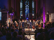 Konzertszene in einer prunkvollen Kirche mit hochaufragenden Fenstern und gotischem Altarraum: Ein Orchester mit Streicher:innen, Bläser:innen und Dirigent verbeugt sich gemeinsam mit einer Sängerin im blauen Abendkleid vor dem applaudierenden Publikum. Bühne und Raum sind in farbiges Licht getaucht.