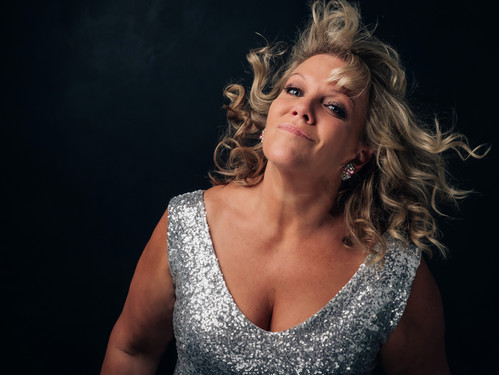 Frau mit lockigem blondem Haar in glitzerndem silbernen Kleid posiert vor dunklem Hintergrund.