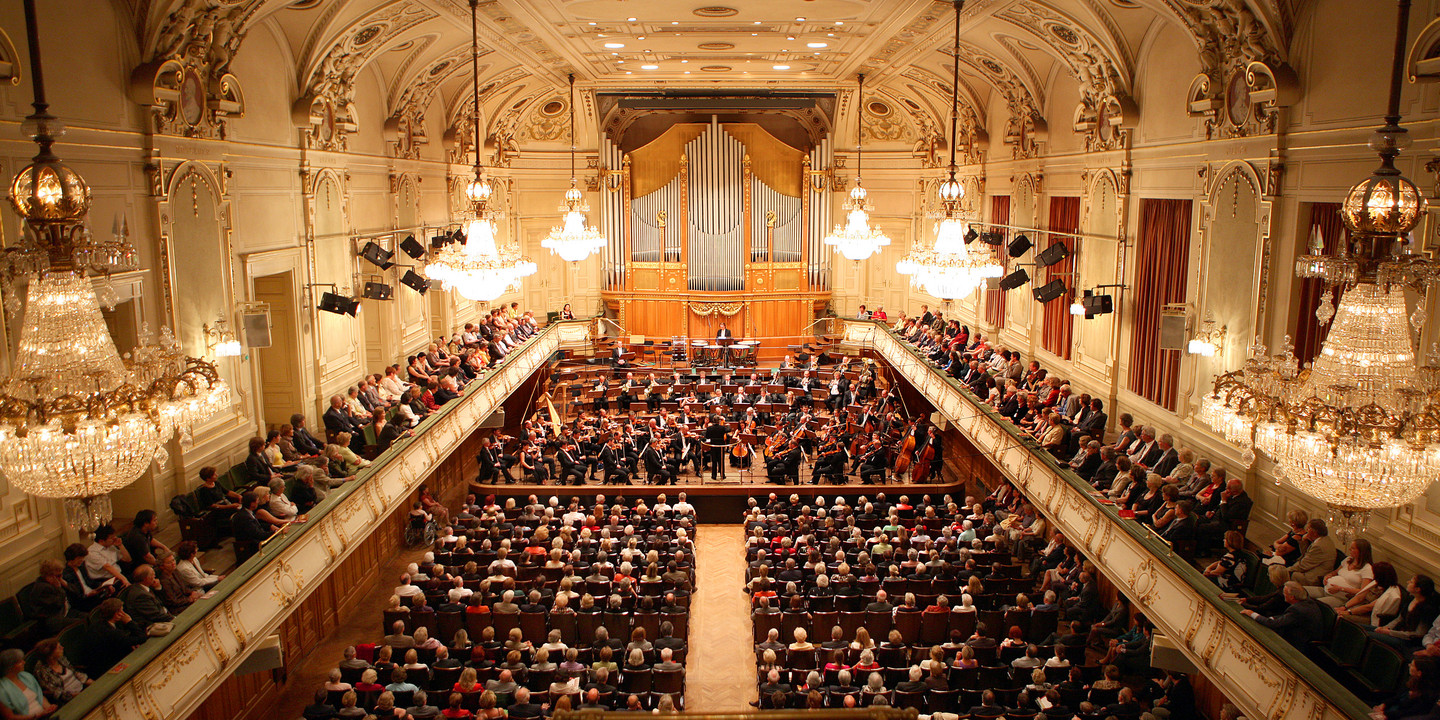 Prunkvoller Konzertsaal mit Publikum, Kronleuchtern und Orchester auf Bühne.